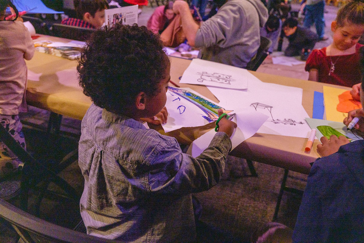 A child at a long, busy table cuts a piece of paper with a colorful drawing on it