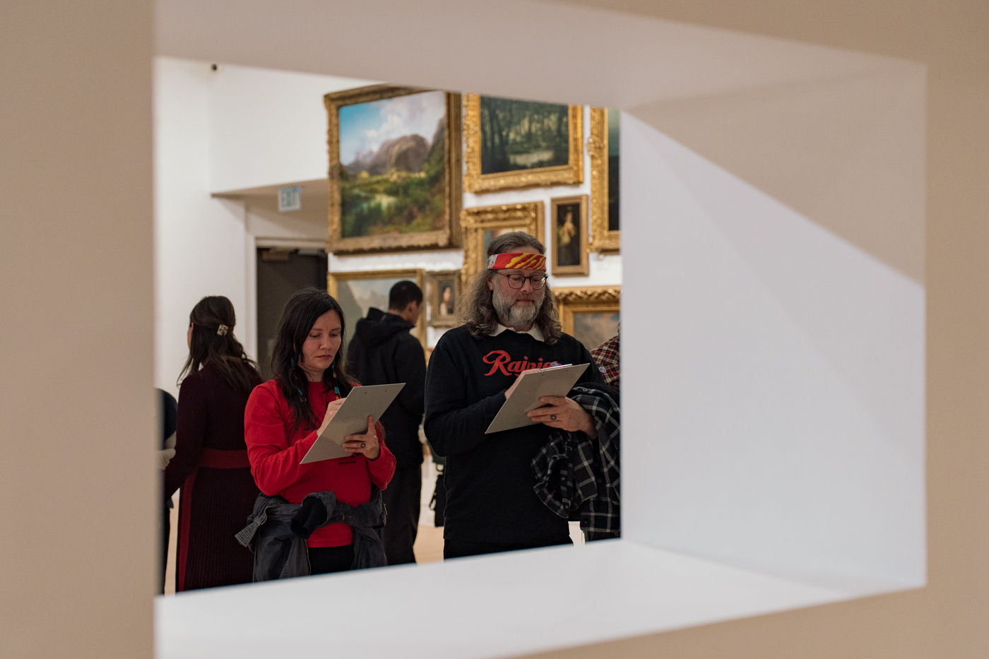People in a gallery holding and writing on clipboards viewed through a square gap in a wall