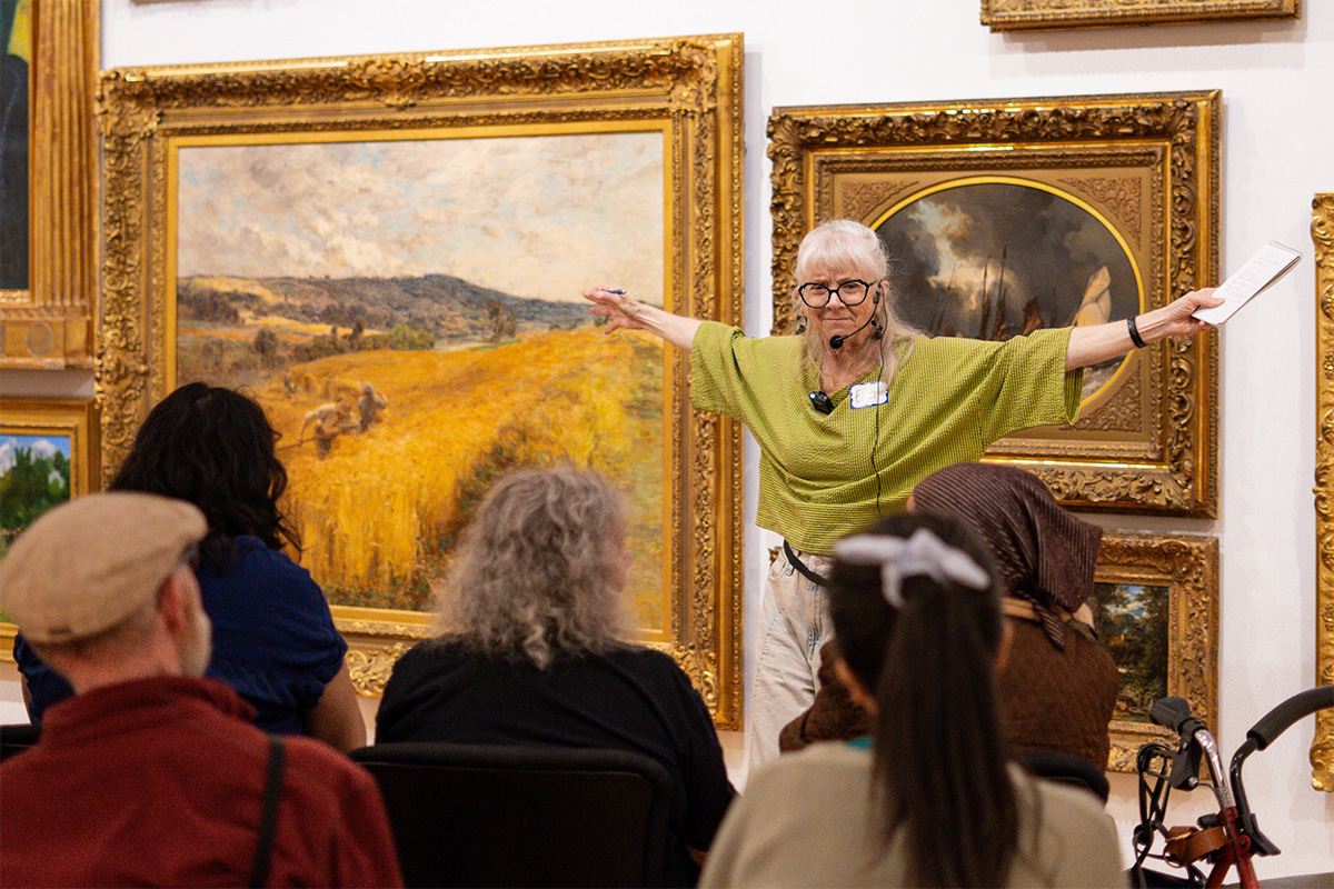 A person holding a notepad with their arms outstretched gesturing and presenting to a seated group of older adults in front of a wall covered in paintings