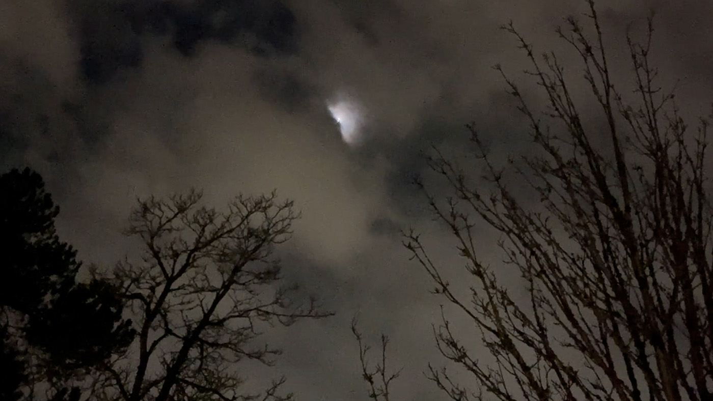 An image of a night sky in an area with trees. The moon is peeking out behind clouds and illuminating the scene