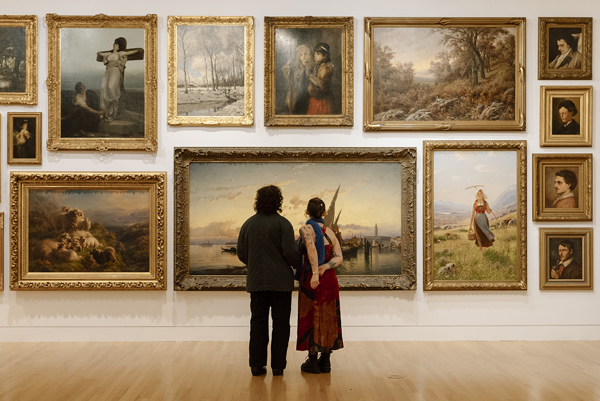 Two people standing in a large room with walls covered in opulently framed oil paintings