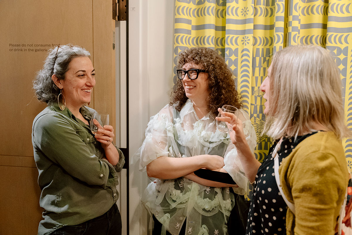 Three people holding drinks and chatting in front of a colorful yellow and blue curtain