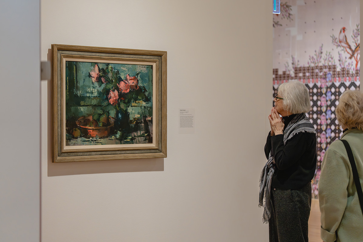 A pair of older women looking at a dark oil painting of a bouquet of flowers hung on a wall. In the background is a glimpse of a colorful, patterned wallpaper