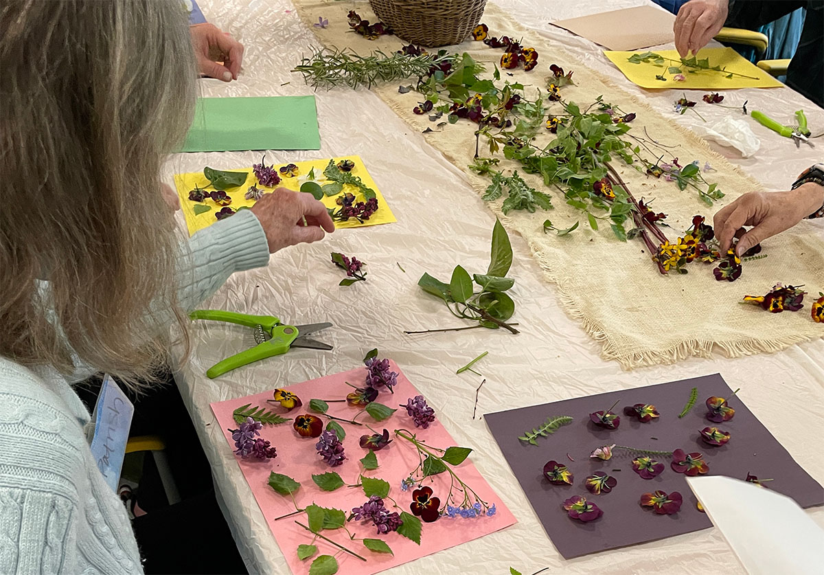 A table covered in papers and flowers for an art project. People's hands are visible as they work and reach for different pieces