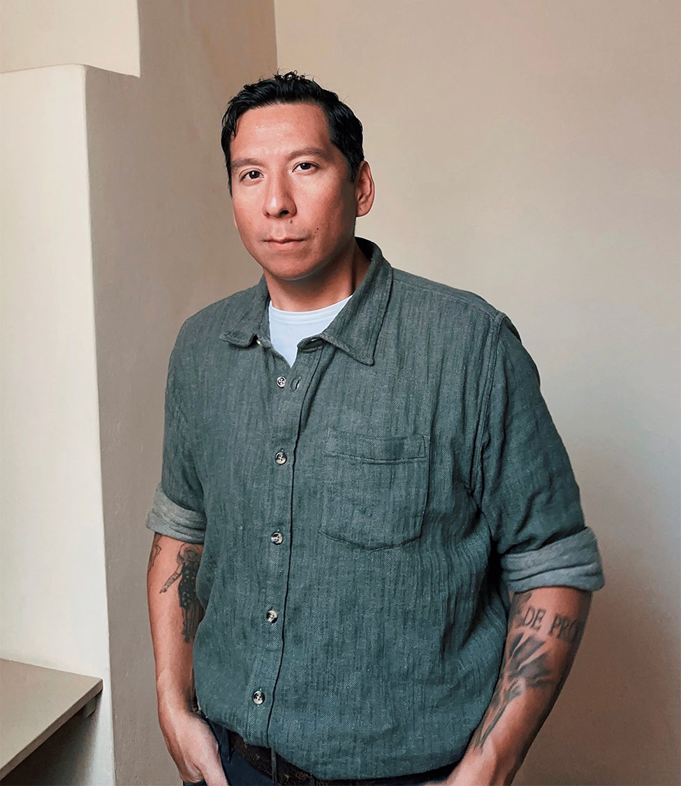 A photo of the artist Sky Hopinka wearing a blue denim button up with the sleeves rolled up