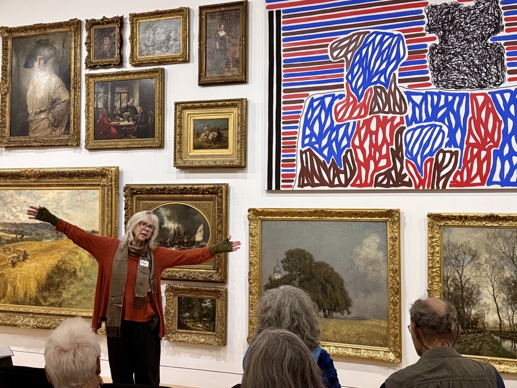 A person leading a class and standing in front of works of art extends their arms for emphasis.