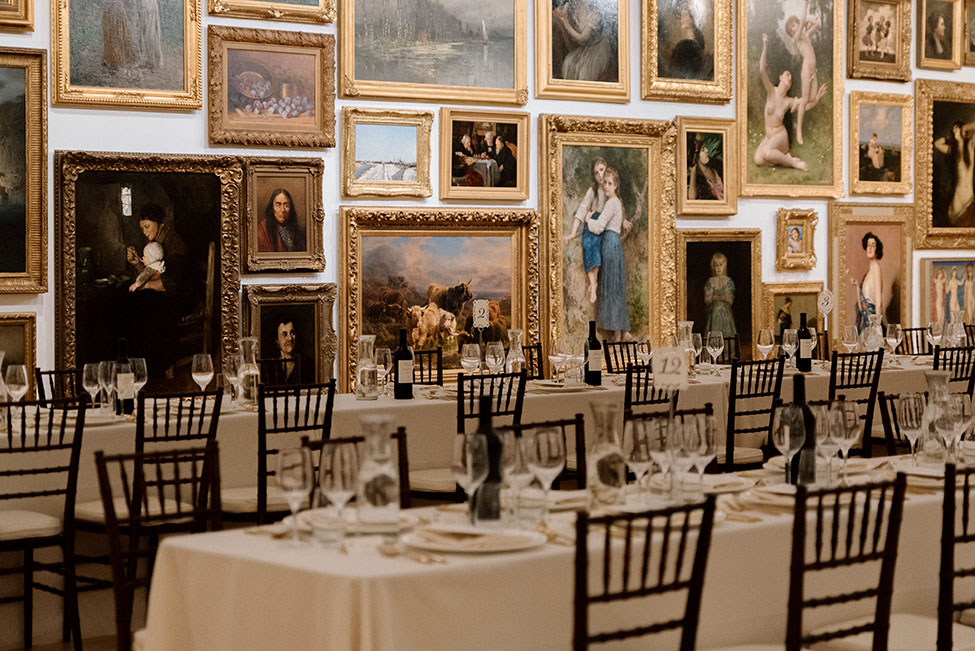 A beautifully set table is captured at the Frye Art Museum, featuring white tablecloths and glassware. The background showcases a gallery wall filled with framed artworks of various styles and subjects.