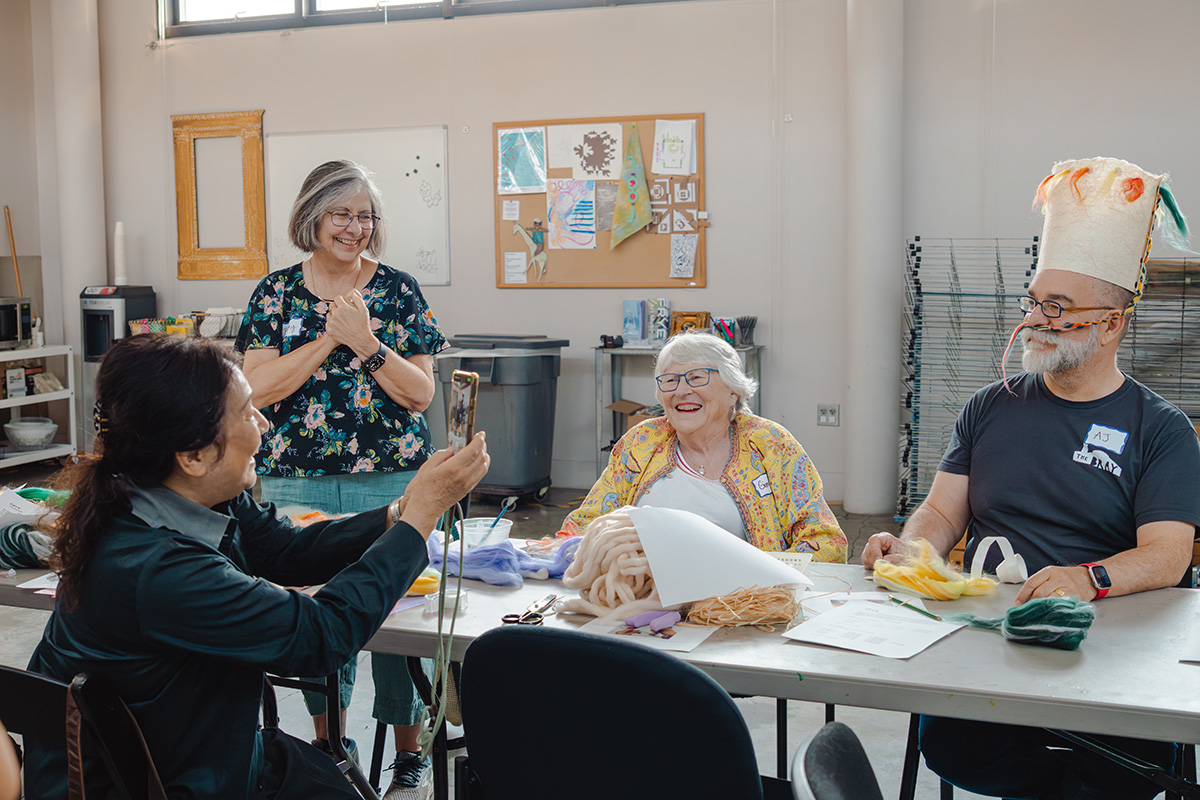 Older adults sitting around a table covered in art materials. A person is taking a photo on their phone of another smiling person.