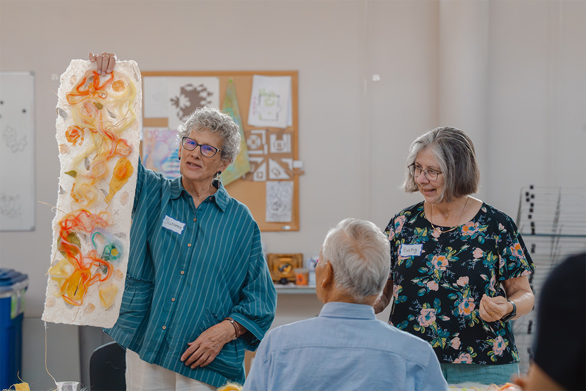 A person holds up a large, rectangular fabric artwork for display to an off-camera group