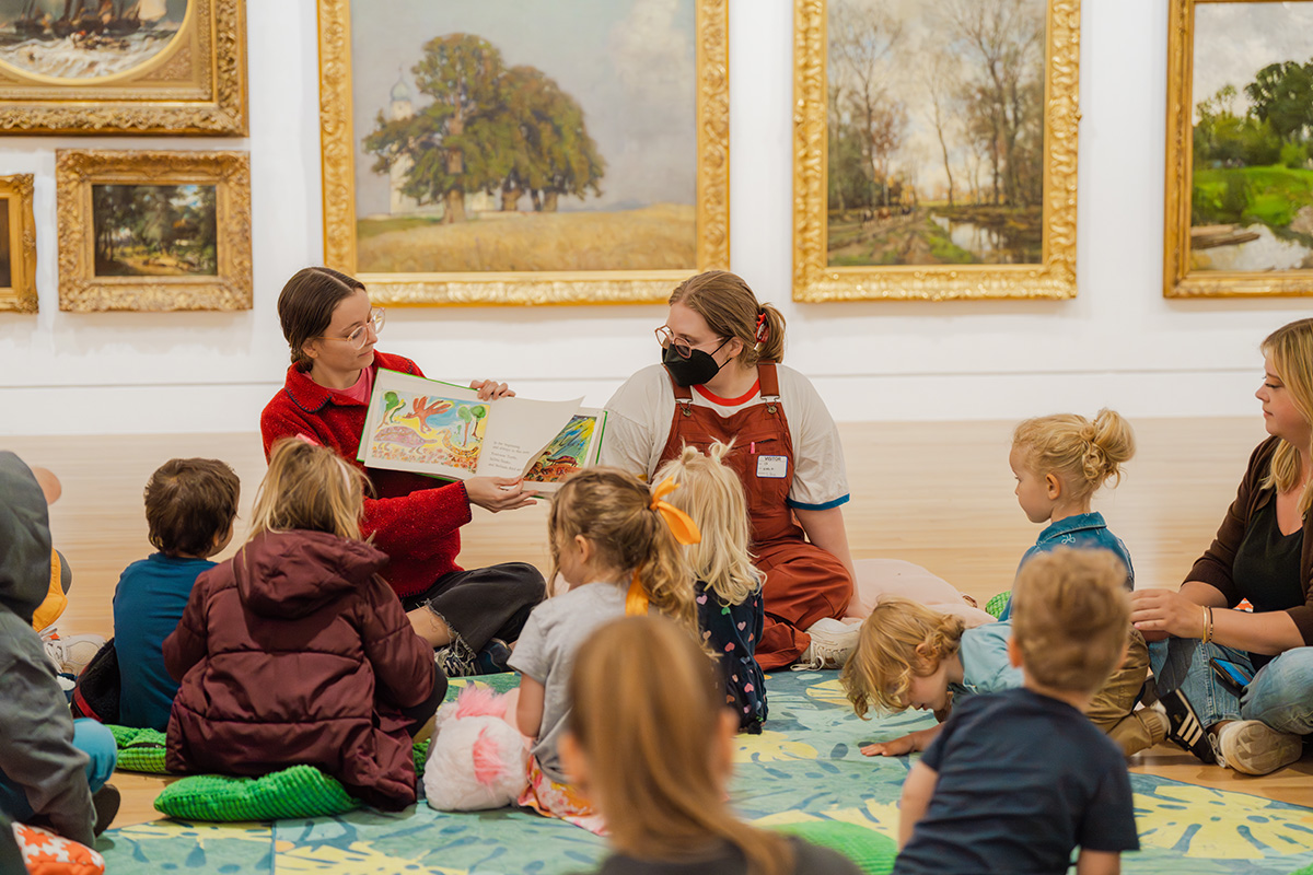 A pair of adults, one holding up a picture book, sitting in front of a group of children in a large art gallery space