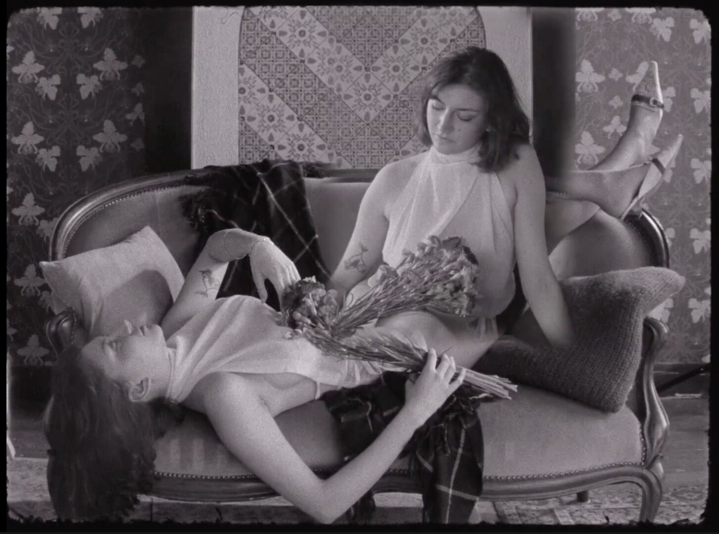 A sepia-toned still from a video of two ghostly female figures entwined on a small loveseat or couch