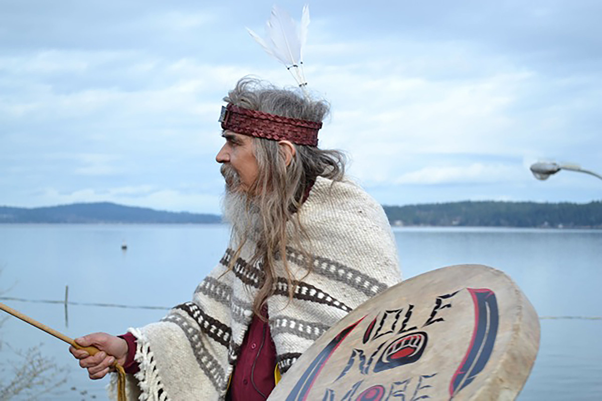 An older man wearing a headband and cloak holding a drum