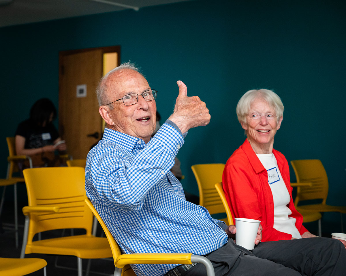An older man and woman seated on yellow chairs. The main is making a thumbs up sign.