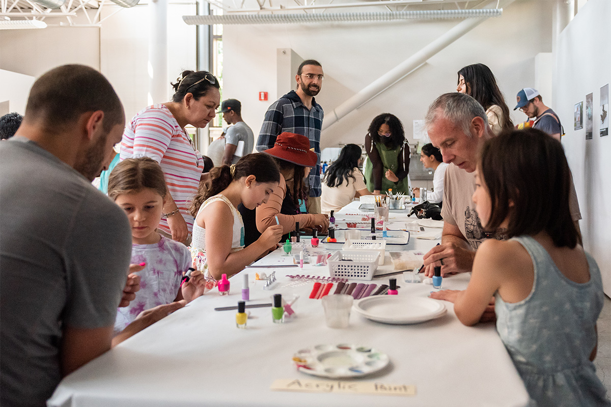Adults and children working on an art activity at a long table covered in a white plastic tablecloth