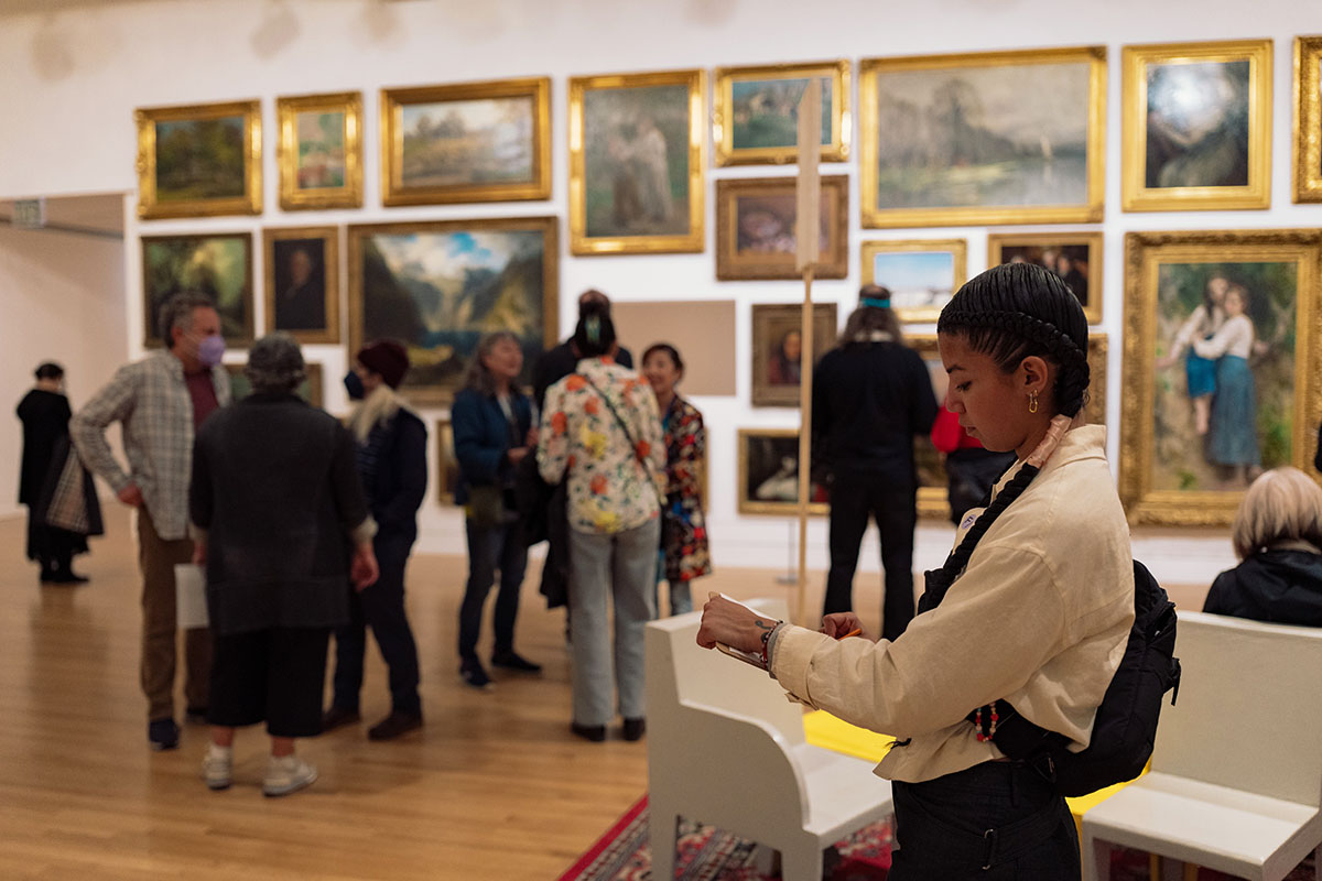 A woman wearing a white shirt and a black bag is writing on a paper on a clipboard while standing in front of a wall of framed landscape paintings in an art  gallery. Several other visitors are gathered around, observing the artwork. The gallery floor is wood, and the room is bathed in soft light.