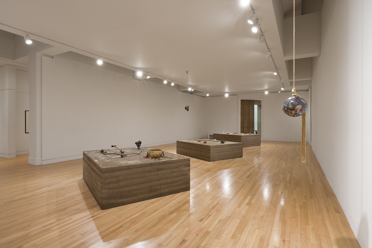 Museum gallery with spherical glass artwork suspended from a rope in the foreground and three pedestals topped with an array of glass artworks in the background