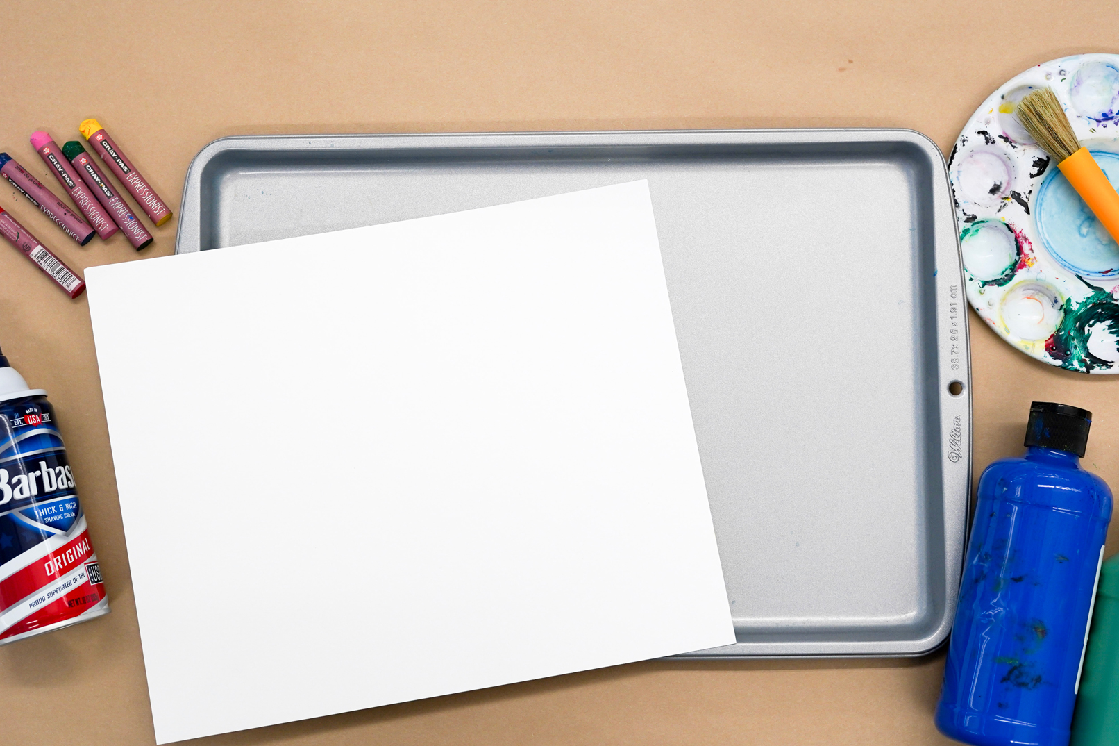 Photo of a blank piece of paper with a sheet pan beneath it and art supplies around including paint, pastels, and shaving cream