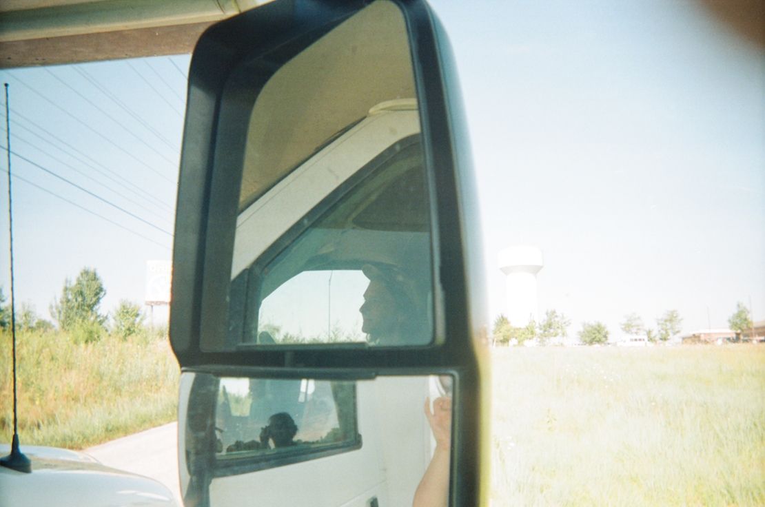 A photograph of a large vehicle's side mirrors with the driver's reflection and landscape in the background