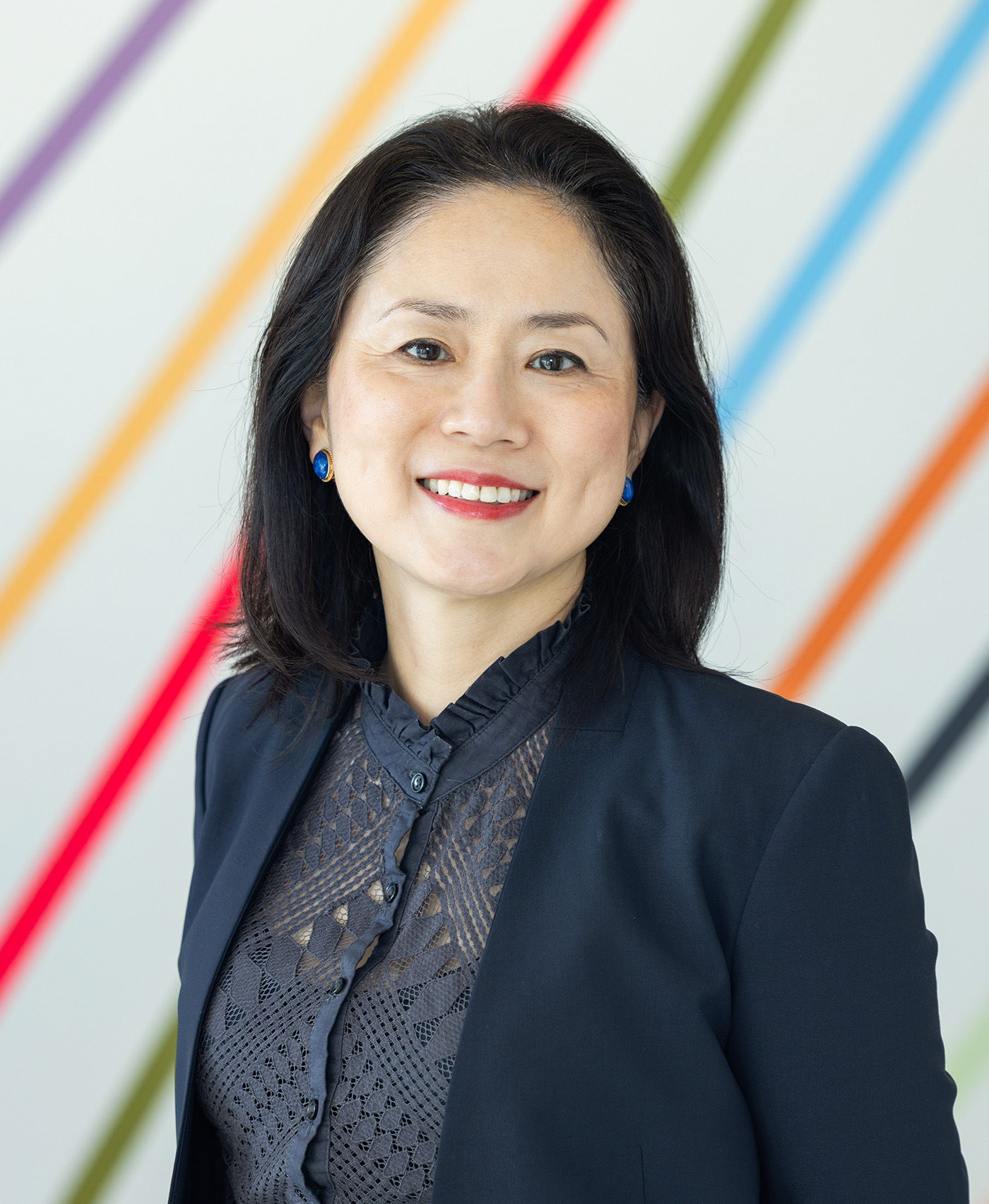 A professional headshot style photo of a smiling woman in formal clothing in front of a wall with colorful diagonal stripes