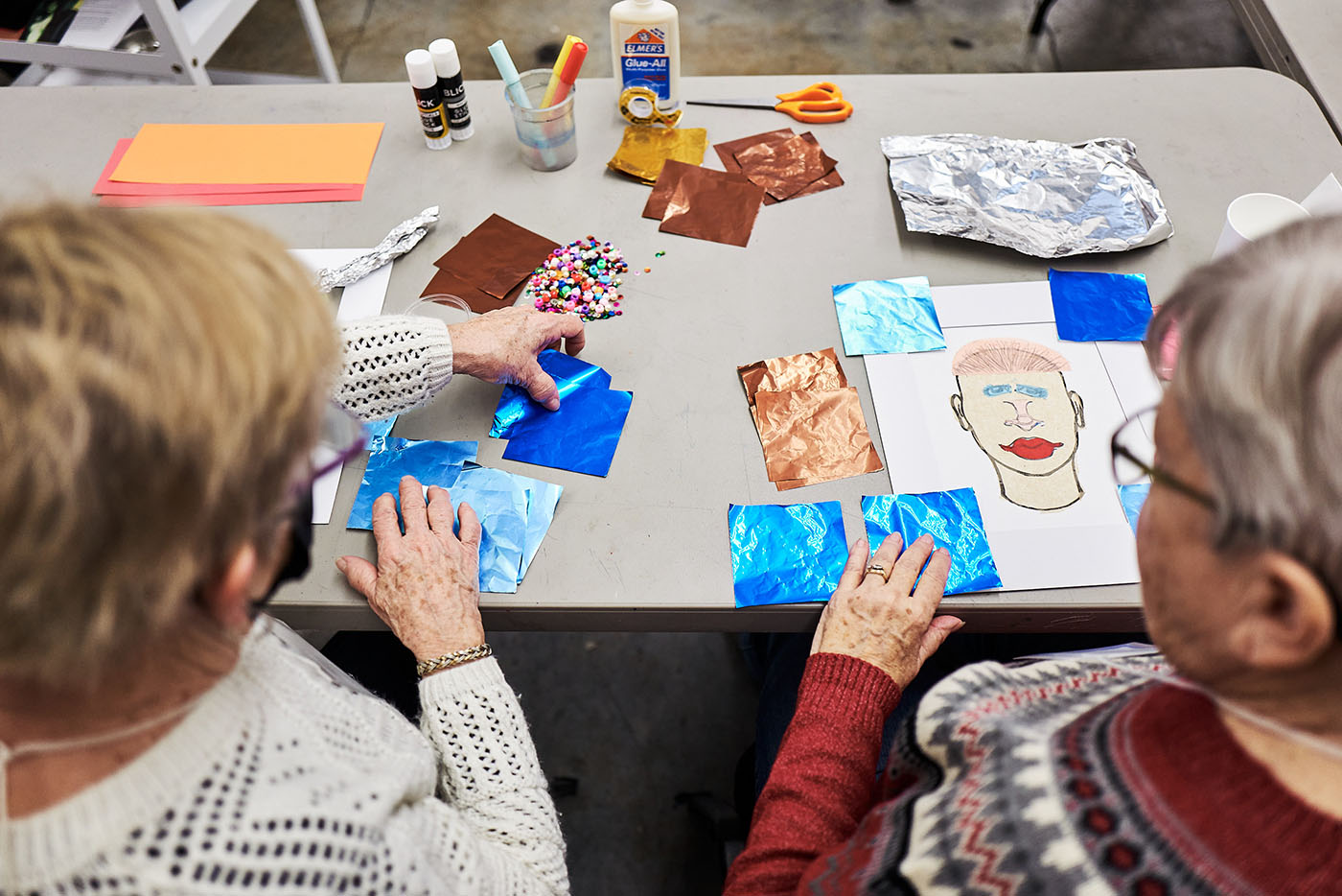 A view from between and behind two older people sitting at a table and working on an arts and crafts project