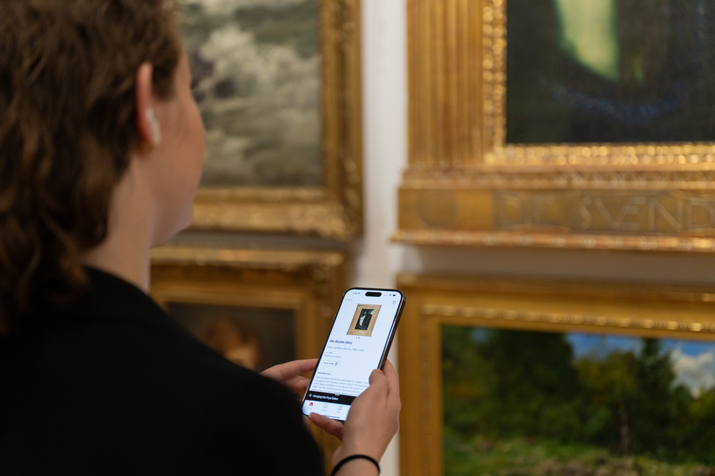A view over a person's shoulder at a phone in their hand. They are standing in front of a wall hung densly with paintings in opulent gold frames.