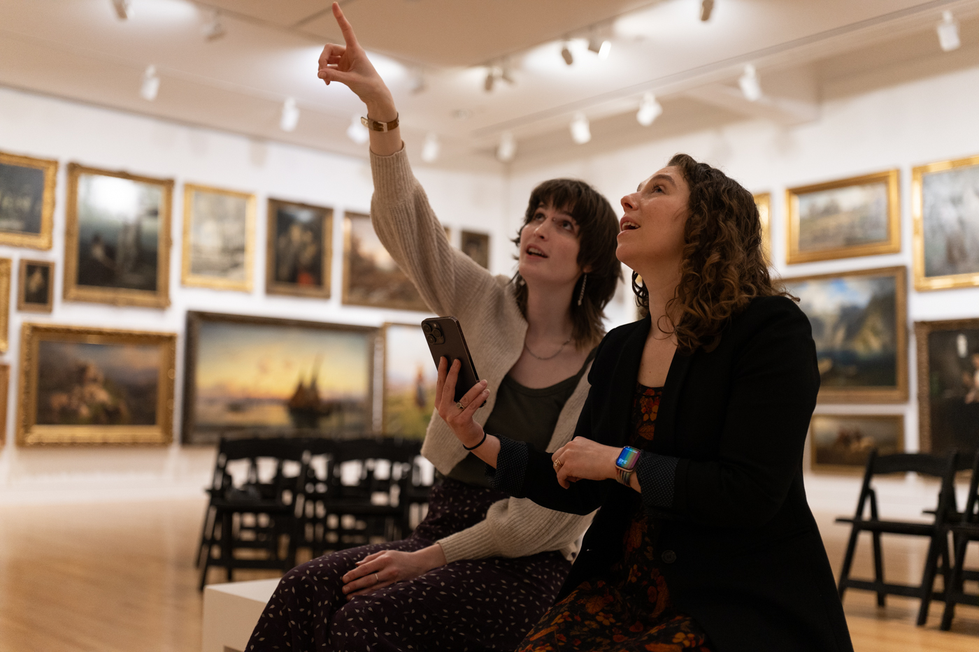 Two women sitting on a bench in a large art gallery space. One is holding a phone for both to be able to view and the other is pointing to something on the wall behind the viewer