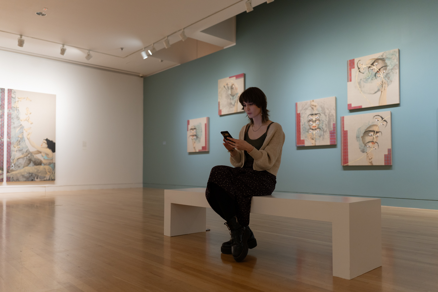 A person sitting on a bench and looking at their phone in a large art gallery. The wall behind them is painted blue and hung with medium-size square paintings