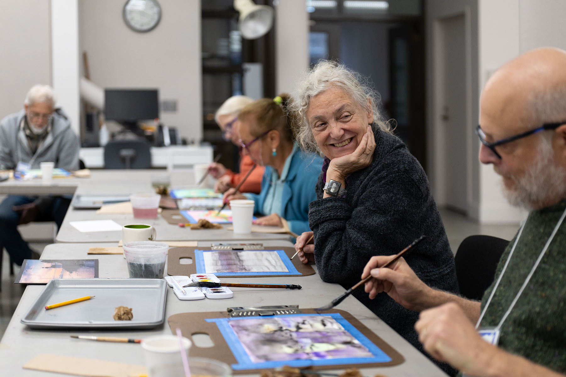 Older adults sitting at a long table and painting with watercolor. One woman is facing the camera and smiling at a person sitting to their left