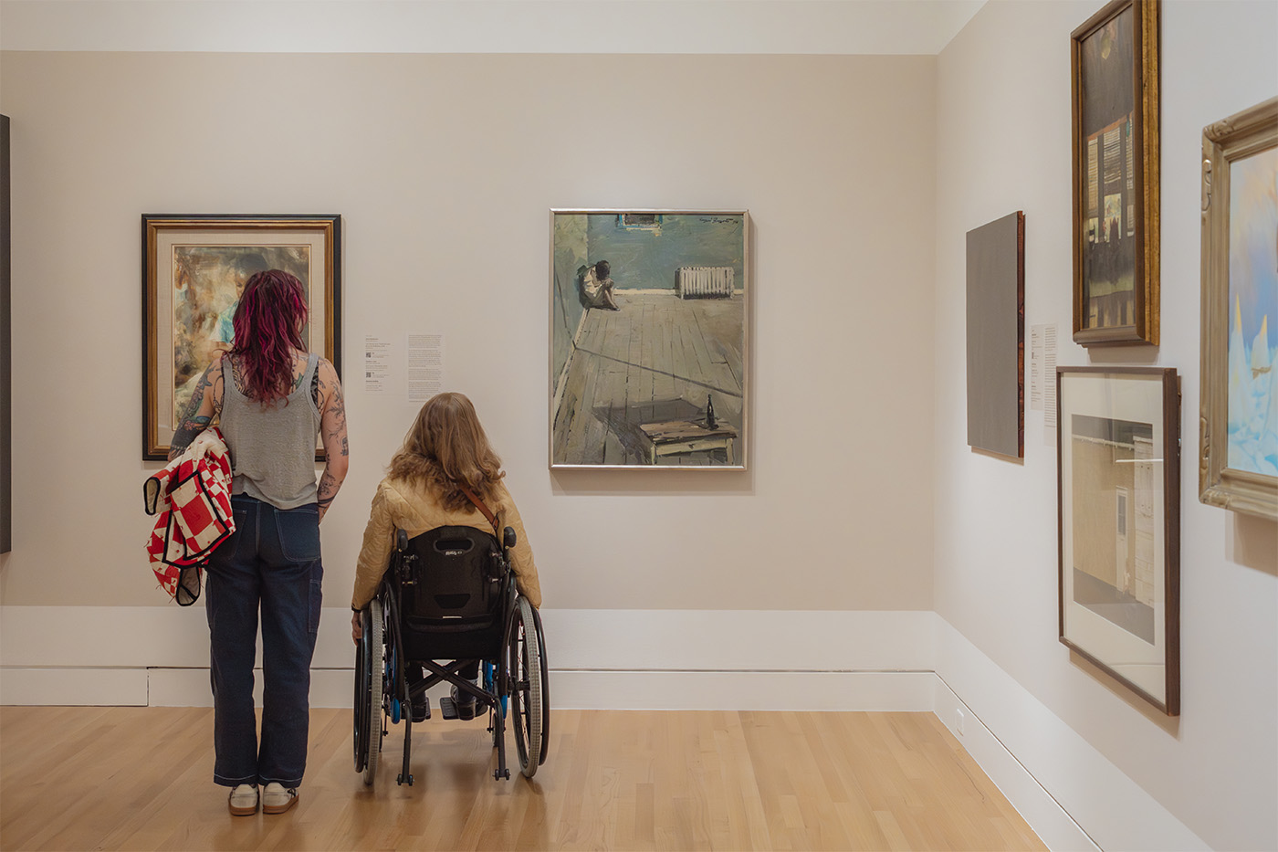 Two people viewed from behind, one standing and one in a wheelchair, both looking at art hanging on a gallery wall