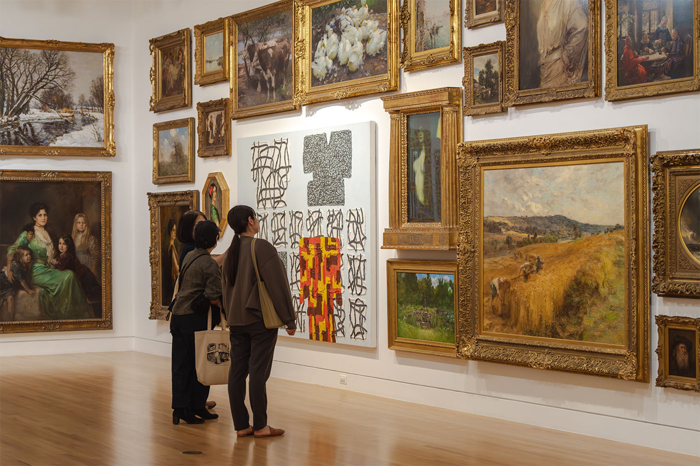 A small group of people standing in a large room decorated in a tight, salon-style hang of paintings