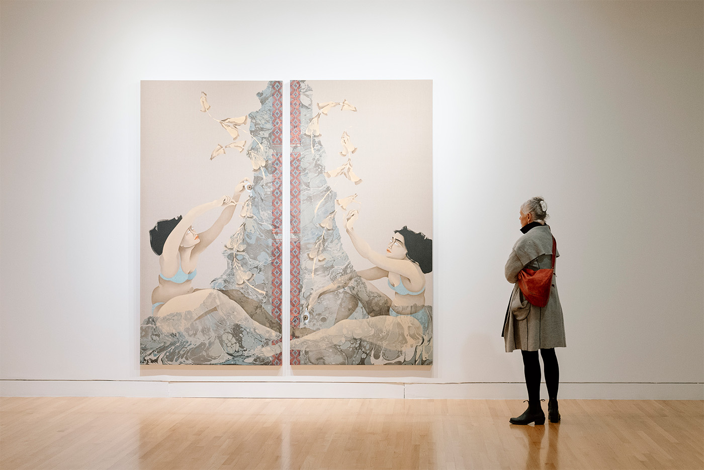 A woman standing to the side and looking at a large painting on canvas of female figures at plant motifs