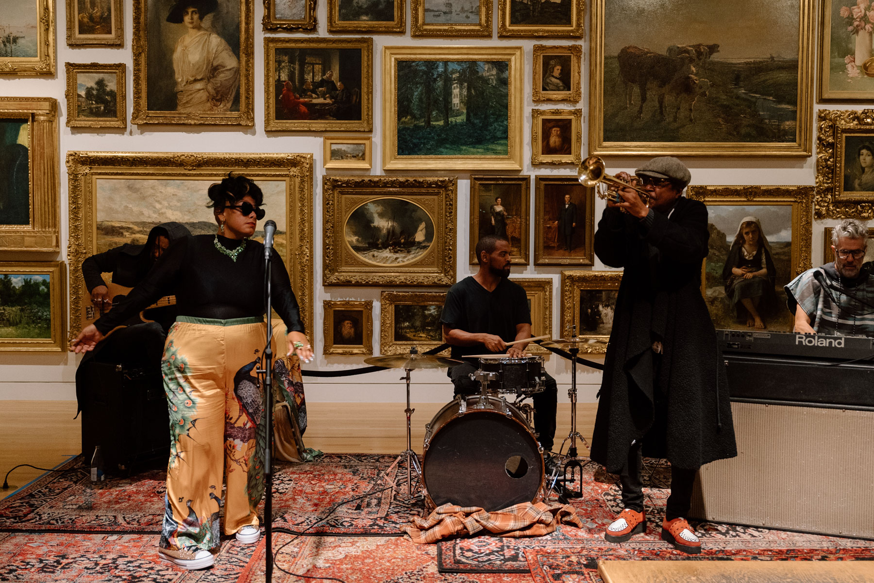 Musicians playing instruments on a colorful rug in front of oil paintings in gold frames hung on a wall