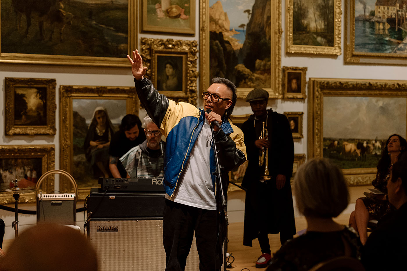 A person performs with a microphone in front of a band, in front of a wall covered in paintings