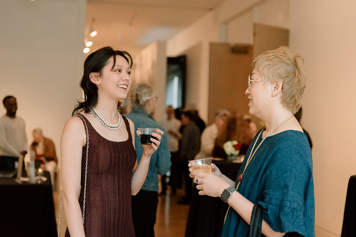 Two women chatting an holding cups of wine in a lightly crowded room