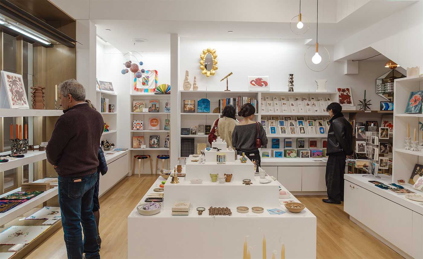 People in a brightly lit museum store space with lots of small items, cards, and books lining the shelves and stacked on pedestals