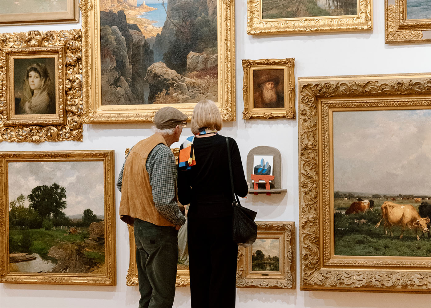 A pair of older people looking at a small sculptural artwork in the midst of a dense arrangement of oil paintings on a wall