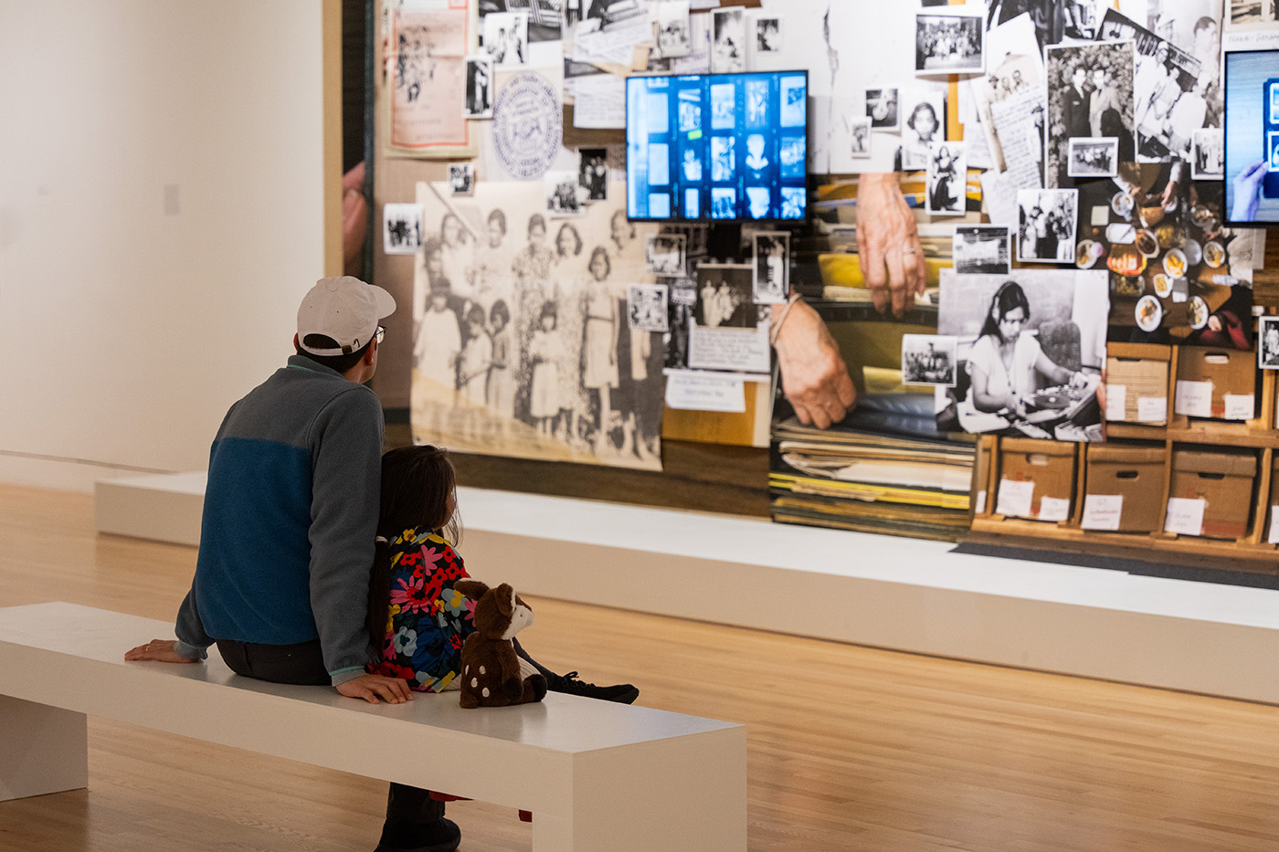 An adult and child sitting together on a white bench in front of a large artwork. The child has a stuffed animal next to them