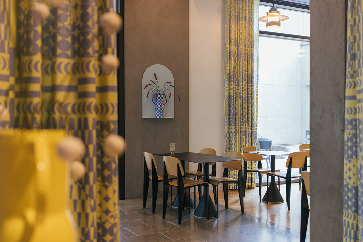 Empty tables in a cafe in front of a large window. There are yellow curtains and an art piece that looks like a flower on the wall