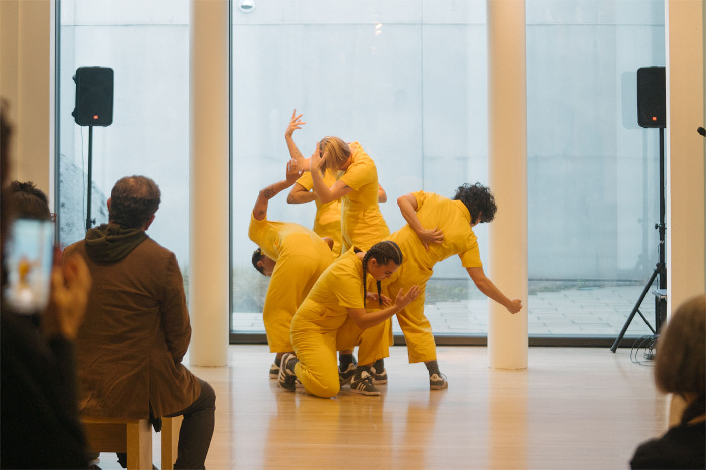 A group of dancers pose in yellow jumpsuits in front of large windows.