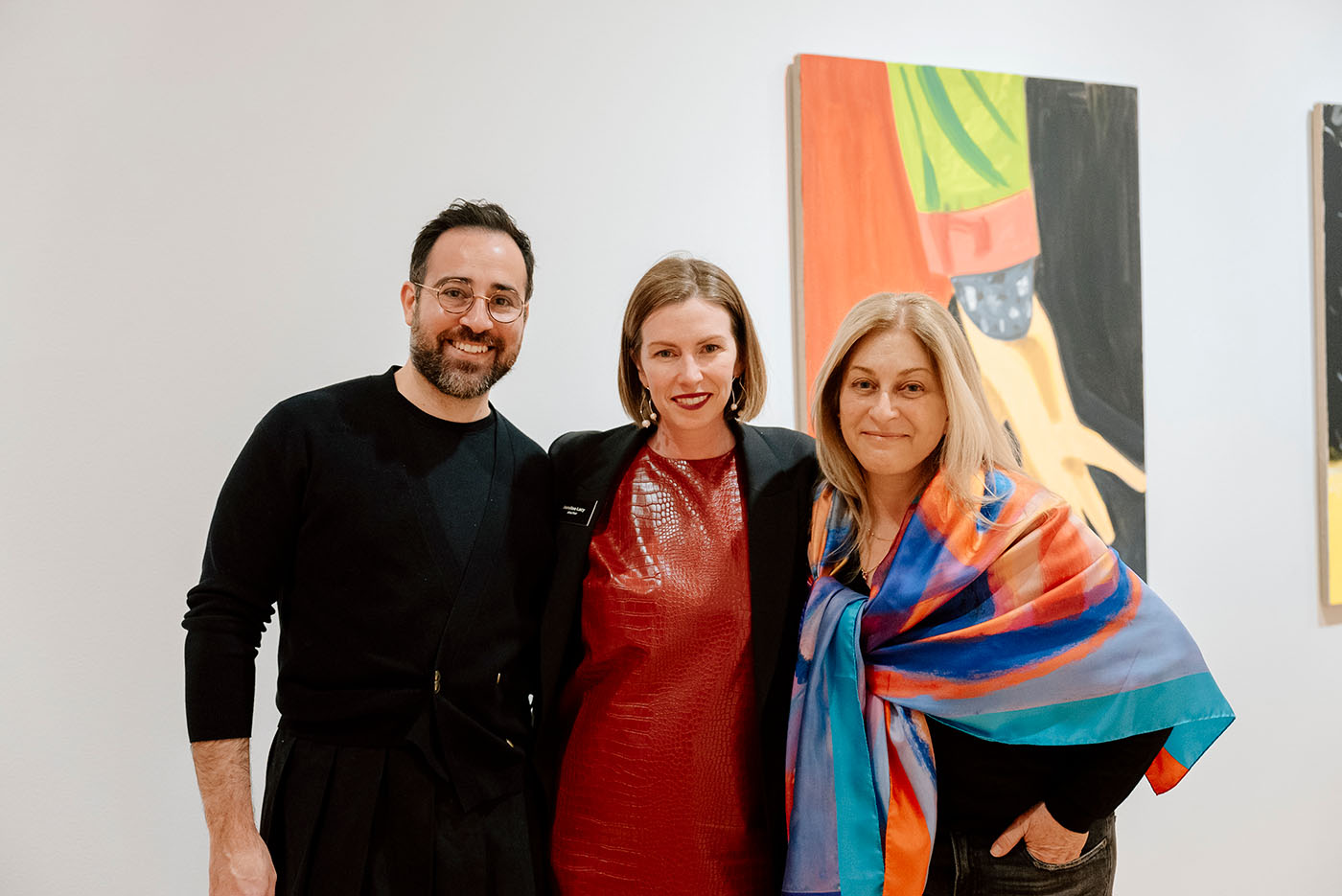 Three people standing closely together and smiling in front of a large, colorful artwork hanging on a wall