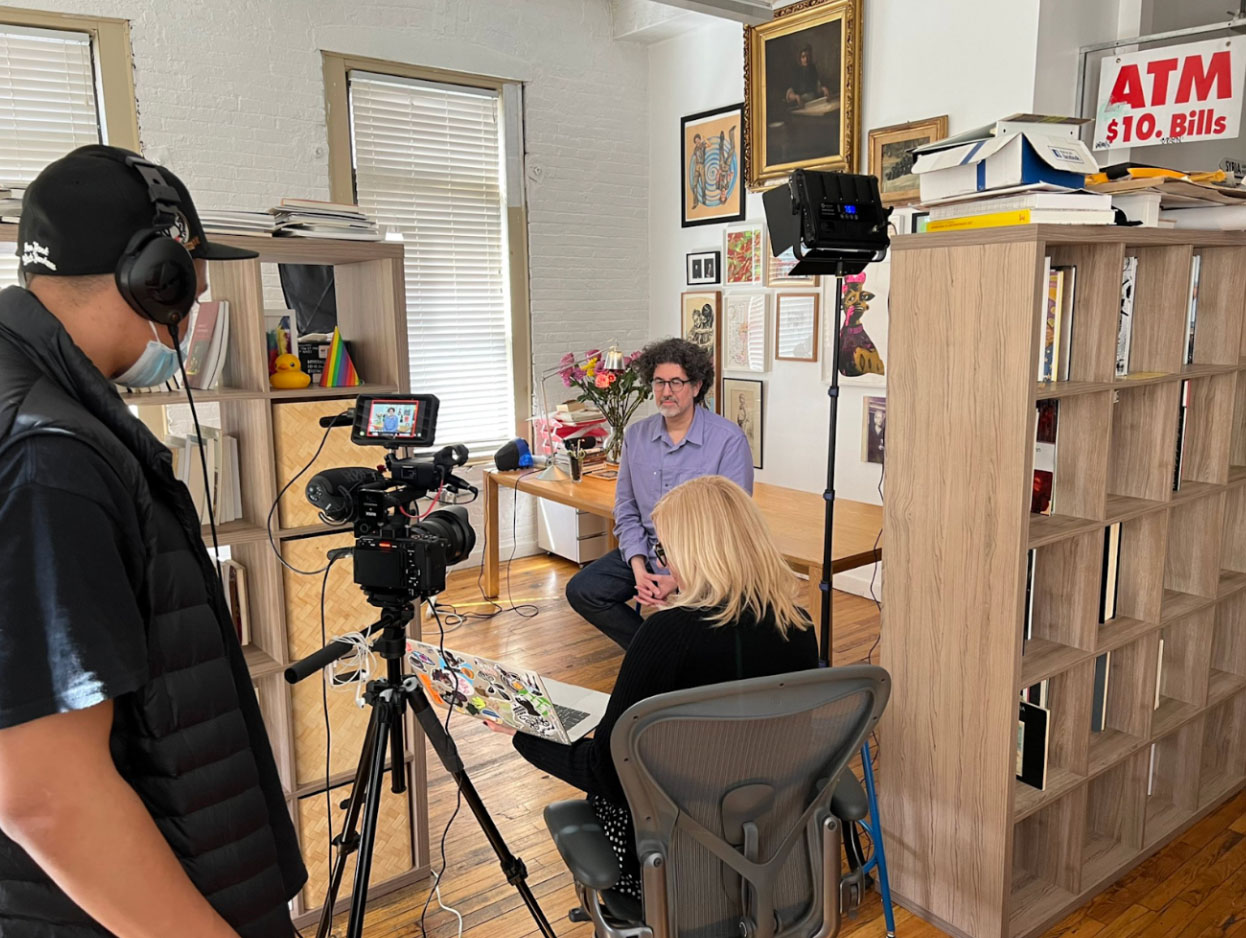 A behind the scenes production photo of a person wearing a blue shirt and sitting on a stool being interviewed on camera. A camera operator and interviewer are also in the frame.