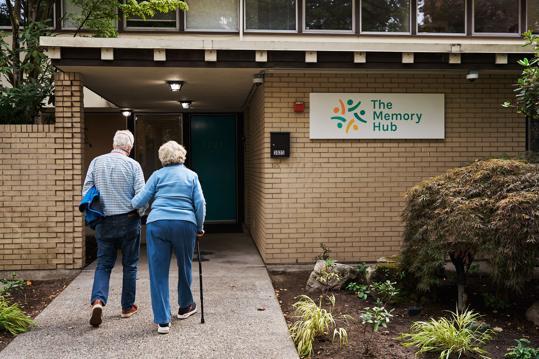 Photo of two people walking down a path leading to a brick building with The Memory Hub sign on the right
