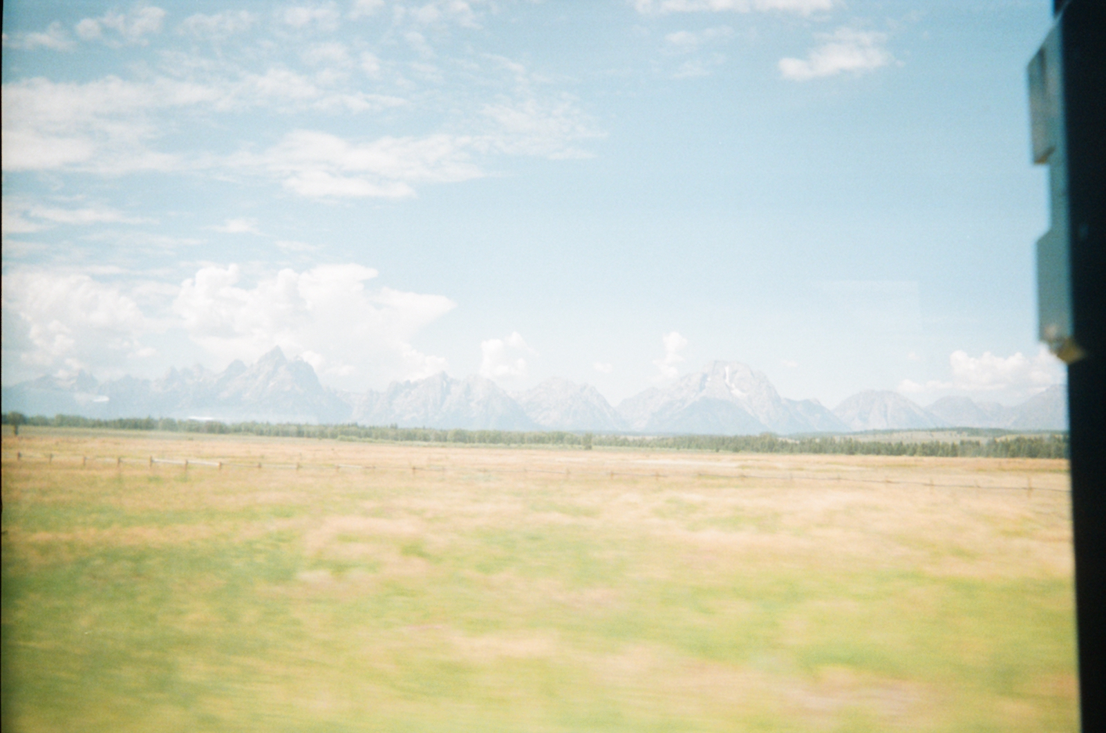 Photograph of far away mountains beyond a green and yellow pasture seen through the window of a moving vehicle