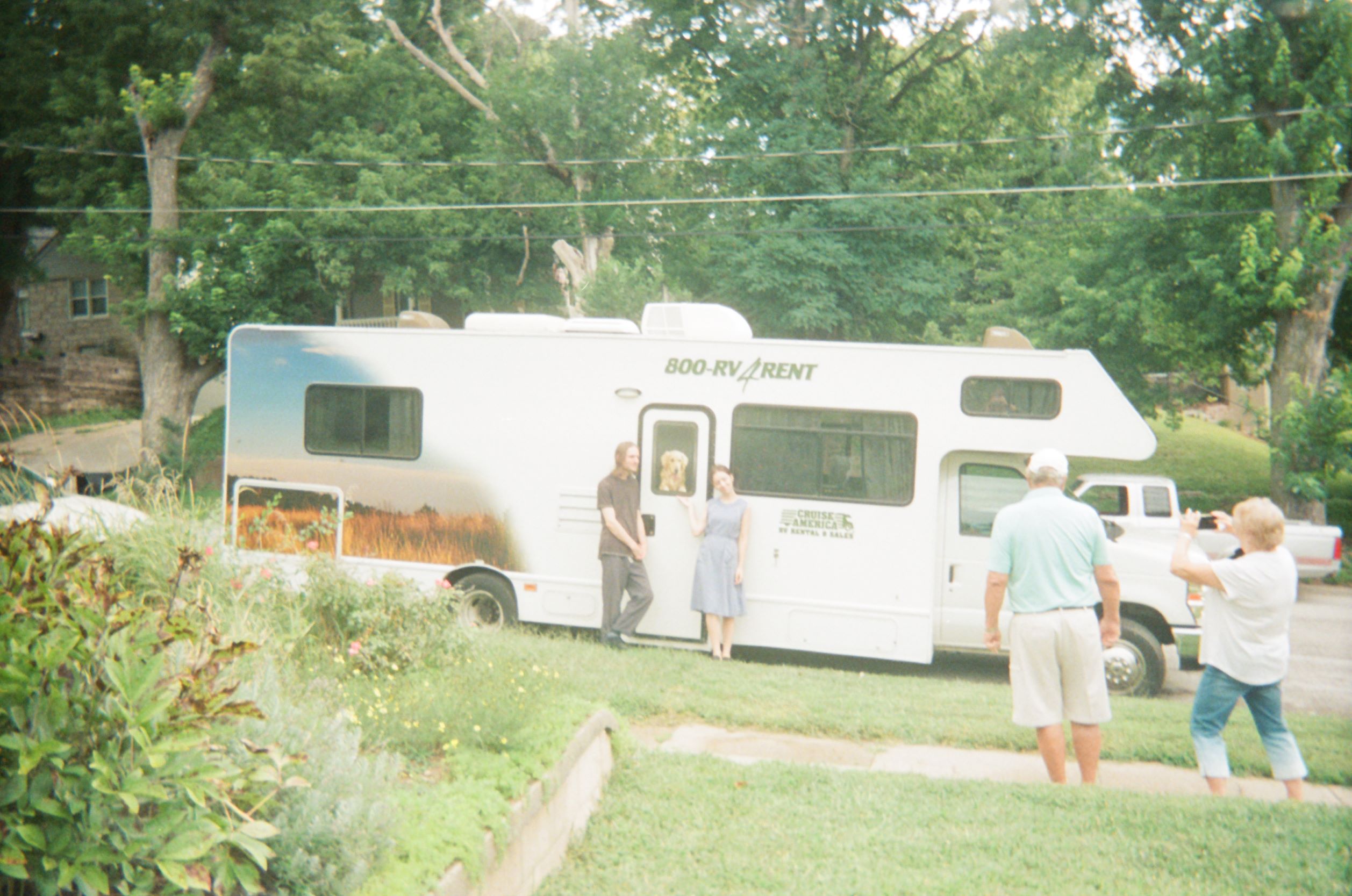 RV in a residential area with two people posing around the door, one person taking a photograph of them, and one person standing nearby