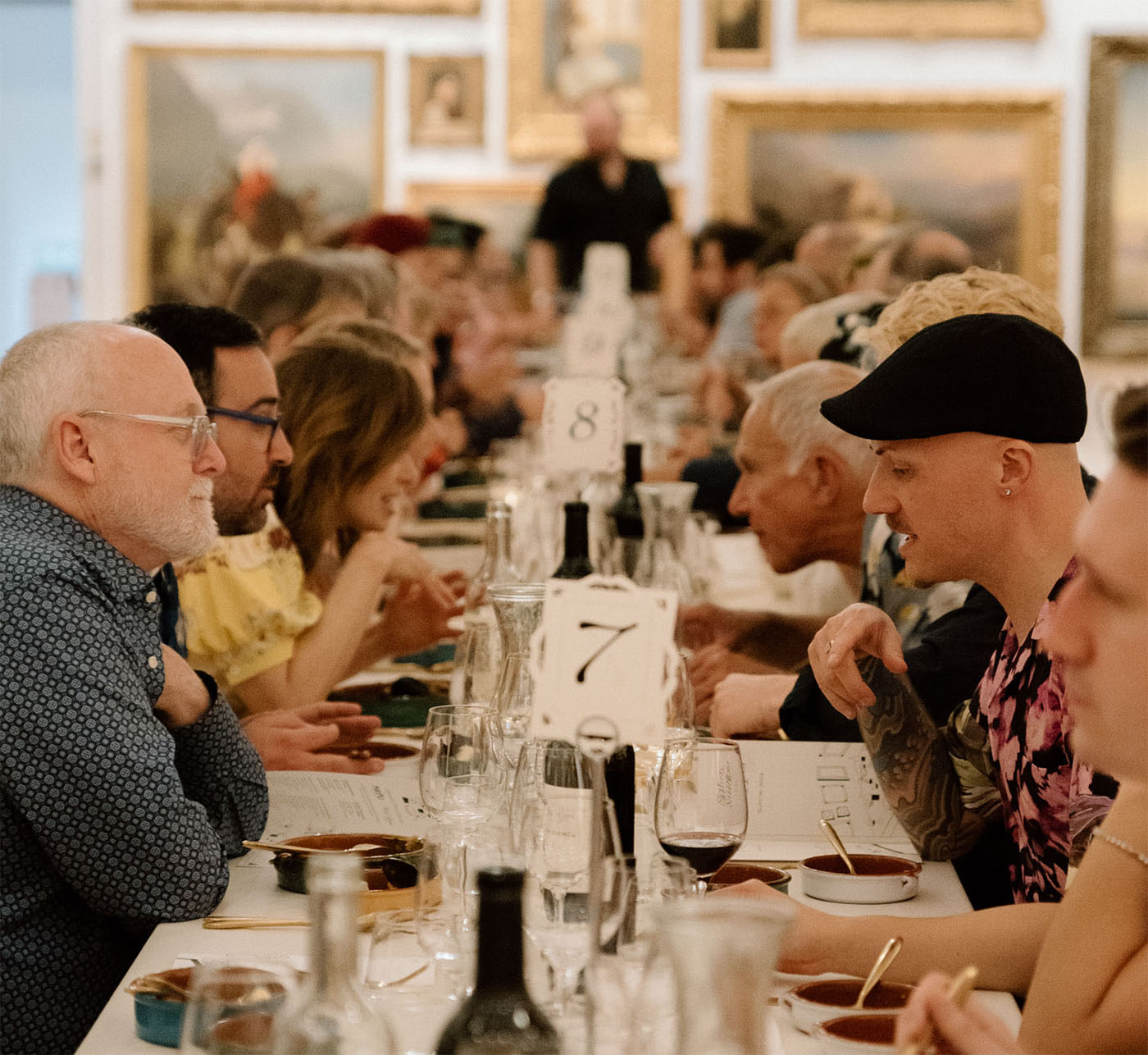 People sitting and talking at a long table laid for dinner in a large gallery with paintings on the walls