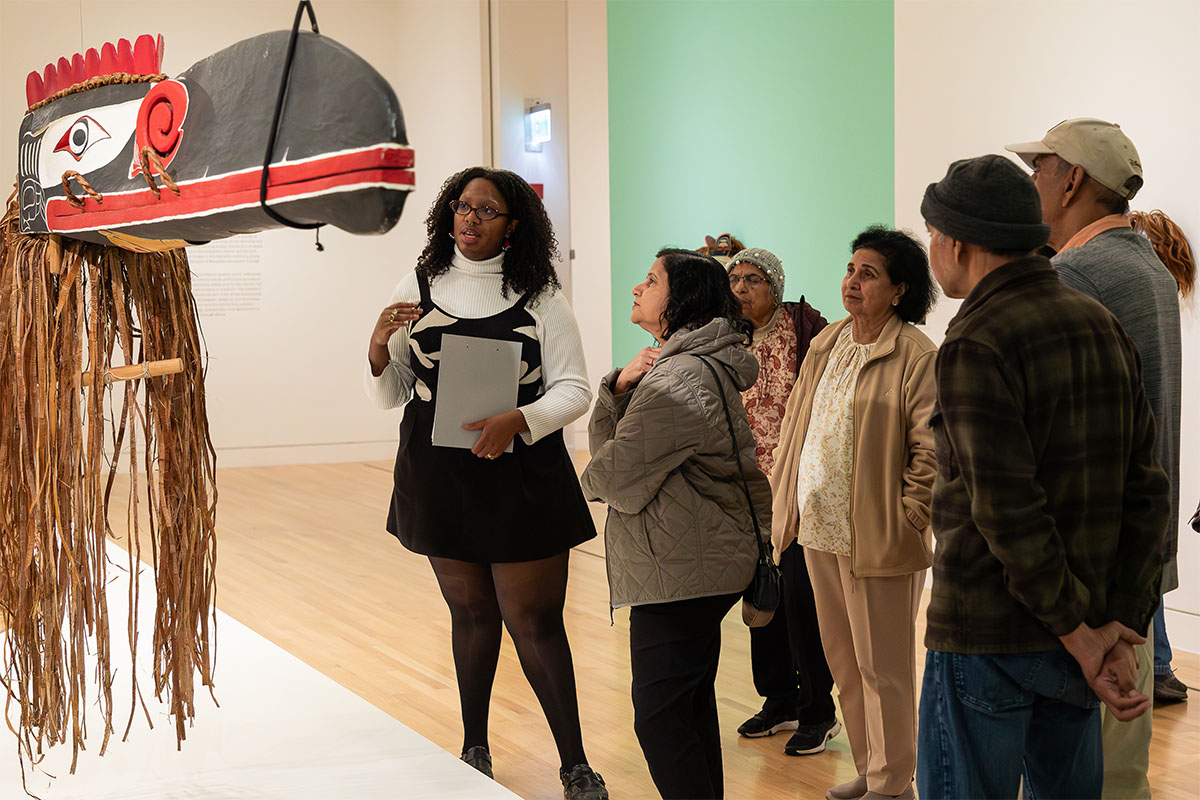 A person leading a group in an art gallery space looking at a large, wooden Pacific Northwest formline sculpture of a raven's head hanging from the ceiling