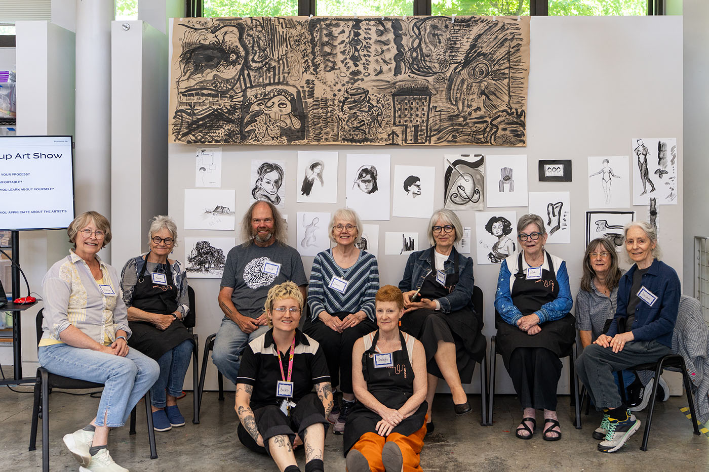 A group of older adults and one younger adult smiling and seated in front of a wall covered in paintings in black ink and watercolor