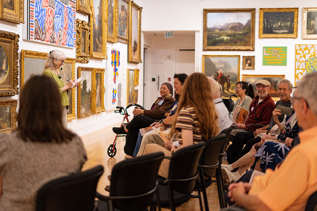 People seated in temporary chairs in an art gallery space facing a presenter standing in front of paintings in gold frames