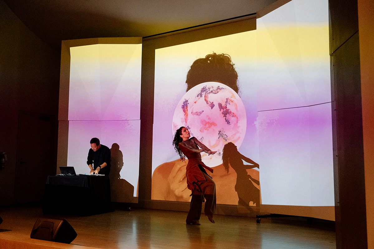 A person dancing on stage in front of an image being project on the wall behind them. Another person to one side of the stage is leaning over electronic music gear on a table.