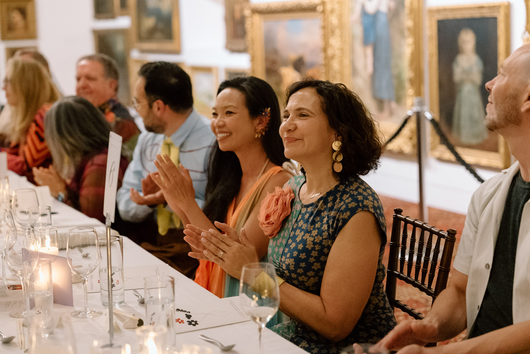 People clapping and seated at a long table laid out for a formal meal in front of a wall covered in oil paintings with opulent gold frames